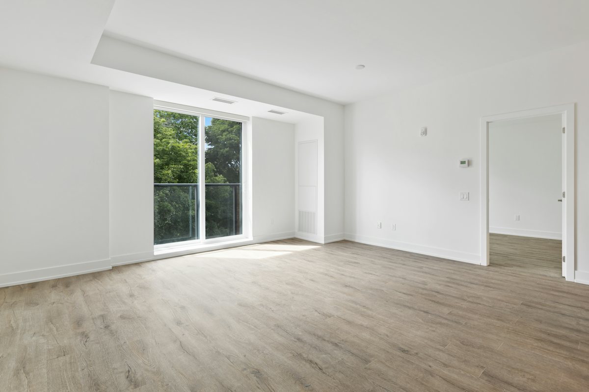 Empty living room with a wide window, white walls, and light wood flooring.